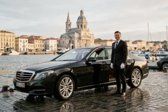 Chauffeur VTC professionnel en costume noir devant une berline de luxe au Vieux-Port de Marseille avec Notre-Dame-de-la-Garde en arrière-plan